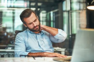 Feeling tired. Young bearded businessman in formal wear massaging his neck while sitting in the modern office. Workplace. Taking a break